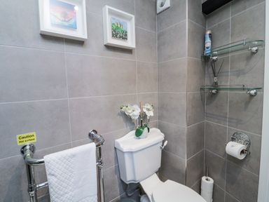 A bathroom with a white toilet a towel on a rack and framed pictures on tiled walls at Nurses Cottage in Alnwick