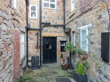 A small stone courtyard with a black door and windows surrounded by stone walls at Nurses Cottage in Alnwick