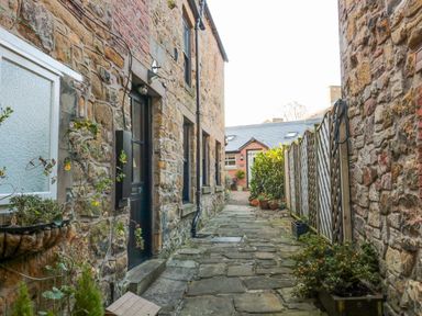 An outdoor stone pathway between stone buildings with plants and a wooden fence at Nurse's Cottage in Alnwick