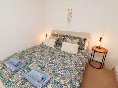A bedroom with floral bedding a small round wooden side table with a lamp and folded towels on the bed at Old Stable Cottage in Alnmouth