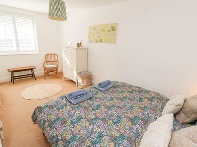 A bedroom with a bed covered in a floral bedspread two blue towels a white dresser a round rug a wicker chair and a bench at Old Stable Cottage in Alnmouth