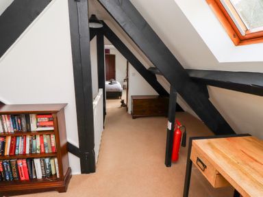 A hallway with black painted beams a bookshelf a wooden desk a fire extinguisher and a view into a bedroom at Old Stable Cottage in Alnmouth
