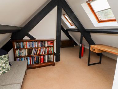 An attic room with a bookshelf filled with books a wooden desk a grey sofa and skylight windows at Old Stable Cottage in Alnmouth