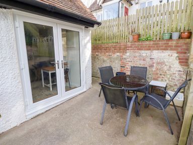 An outdoor patio area with a round glass table and four black chairs next to white double doors and a brick wall with potted plants at Old Stable Cottage in Alnmouth