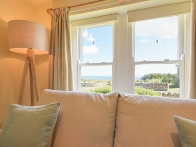 A beige sofa with green cushions near a window with a view of a garden and sea and a floor lamp at Ottoline Cottage in Embleton