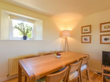 A dining room with a wooden table and chairs a bowl of fruit on the table a floor lamp in the corner and framed pictures on the wall at Ottoline Cottage in Embleton