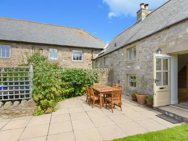 A stone patio with a wooden table and six chairs near a stone building with plants in pots at Ottoline Cottage in Embleton