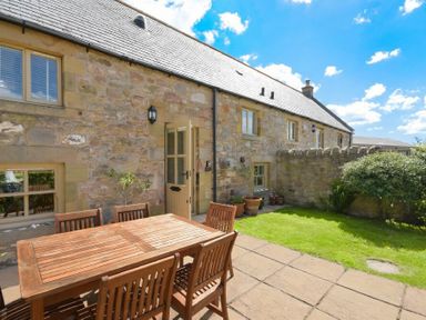 An outdoor patio area with a wooden dining table and chairs next to a stone wall house with potted plants and grass at Ottoline Cottage in Embleton
