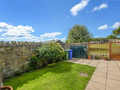 A small garden with grass next to a stone wall and a wooden gate with bins at Ottoline Cottage in Embleton