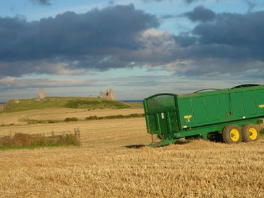 A green farm trailer in a harvested field with ruins on a hill in the distance at Ottoline Cottage in Embleton