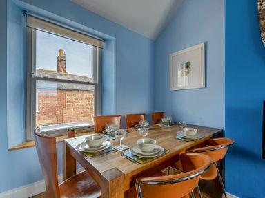 A dining room with a wooden table set for six and brown chairs by a window at Pilots Rest in Alnmouth