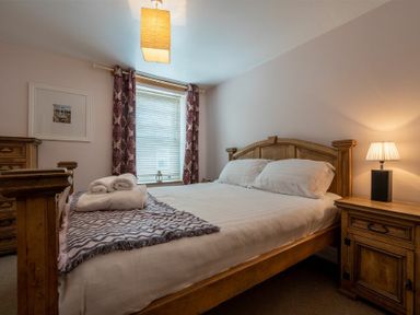 A bedroom with a wooden bed, side table with lamp, chest of drawers, window with curtains and folded towels on the bed at Pilots Rest in Alnmouth