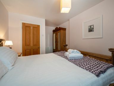 A bedroom with a wooden bed frame white bedding rolled towels on a blanket a wooden wardrobe a nightstand with a lamp and a closed wooden door at Pilots Rest in Alnmouth