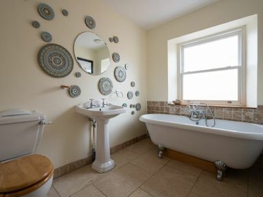 A bathroom with a toilet with wooden seat a pedestal sink a bathtub under a window and a round mirror on the wall at Pilots Rest in Alnmouth