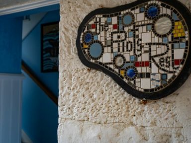 A mosaic artwork on a textured stone wall with a blue room and staircase in the background at Pilots Rest in Alnmouth