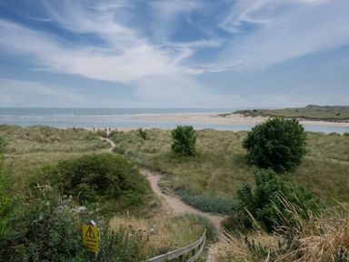 A sandy path through dunes and bushes leading to a beach and sea under a partly cloudy sky at Pilots Rest in Alnmouth