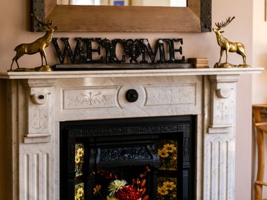 A fireplace with decorative details and floral tiles with a welcome sign and two golden deer statues on the mantel at Upper Alnbank in Alnmouth