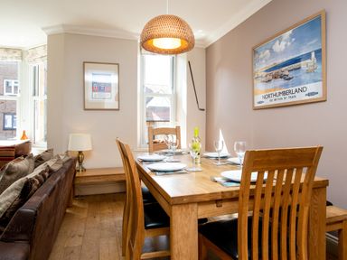 A dining area with a wooden table set with plates and glasses and chairs in a room with framed pictures at Upper Alnbank in Alnmouth