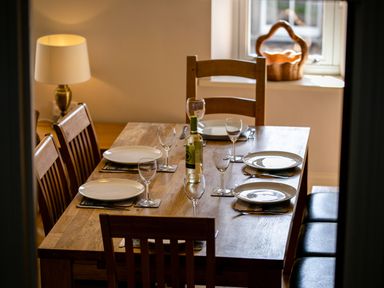A wooden dining table set with plates and wine glasses with a bottle of white wine and a lamp in the background at Upper Alnbank in Alnmouth