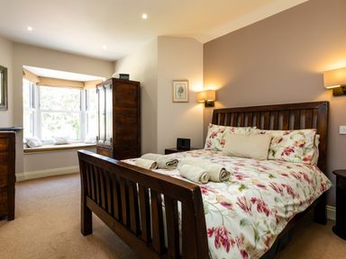 A bedroom with a wooden bed with floral bedding and rolled towels with a wooden wardrobe and window seat at Upper Alnbank in Alnmouth