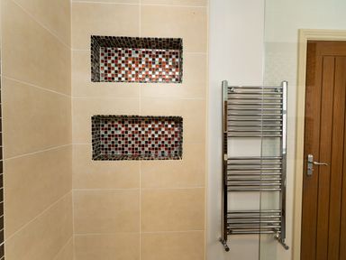 A bathroom wall with built in shower shelves tiled with small red and gray mosaic tiles and a chrome heated towel rail beside a wooden door at Upper Alnbank in Alnmouth