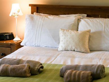 A made bed with white pillows and beige decorative cushion beside a bedside table with lamp and folded towels at Upper Alnbank in Alnmouth