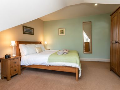 A bedroom with a wooden bed with white bedding and green throw a wooden nightstand with a lamp a tall mirror on the wall and a wooden wardrobe at Upper Alnbank in Alnmouth