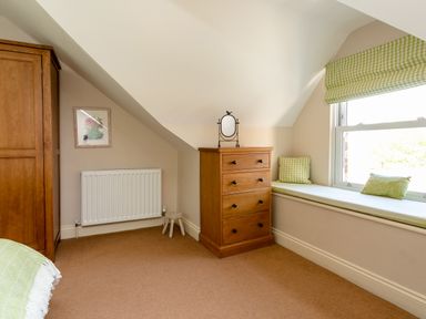 A bedroom corner with a wooden chest of drawers a small mirror a window seat with cushions and a wooden wardrobe at Upper Alnbank in Alnmouth