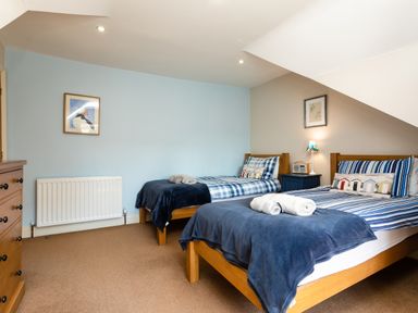 A bedroom with two single beds with blue blankets and striped pillows a wooden dresser and a framed puffin picture on a blue wall at Upper Alnbank in Alnmouth