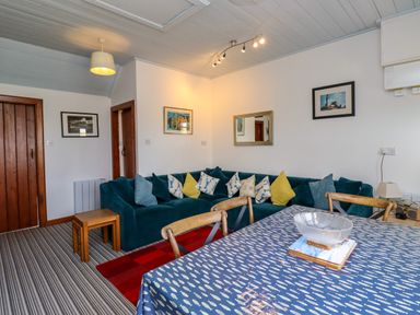 A living room with a blue corner sofa and multiple cushions a dining table with a patterned tablecloth and wooden chairs at Sand Dune Cottage in Bamburgh