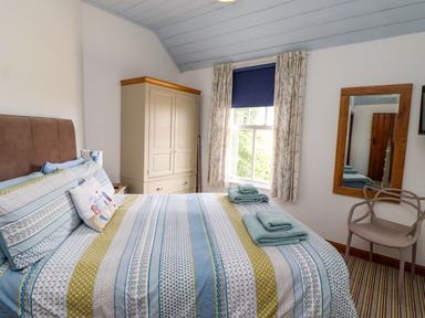 A bedroom with a bed towels on the bed a wardrobe a window with curtains a wall mirror and a chair at Sand Dune Cottage in Bamburgh