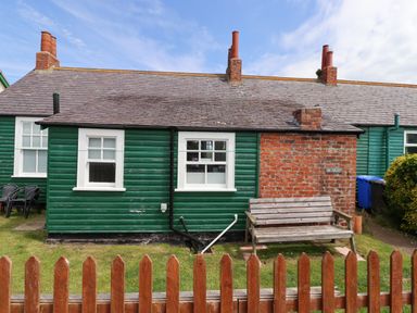A green wooden cottage with white windows a wooden bench and a small front yard with a fence at Sand Dune Cottage in Bamburgh