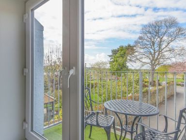 A balcony with a round metal table and chairs overlooking trees and a road at Schooner Court No1 in Alnmouth
