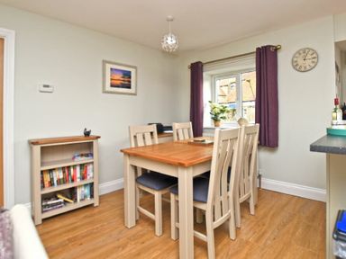 A dining area with a wooden table and chairs near a window with purple curtains and a small bookshelf at Schooner Court No1 in Alnmouth