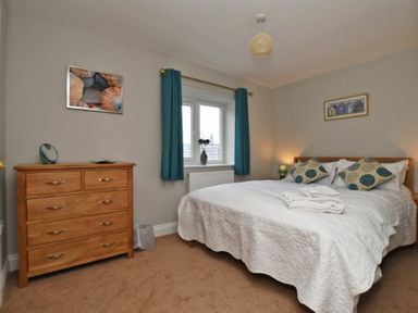 A bedroom with a double bed with white bedding two patterned pillows a wooden dresser a window with blue curtains and framed pictures at Schooner Court No1 in Alnmouth