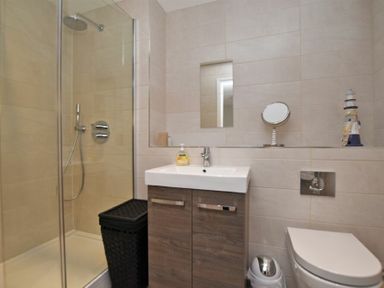 A bathroom with a shower enclosure a sink with cabinet a toilet a mirror and a black laundry basket at Schooner Court No1 in Alnmouth