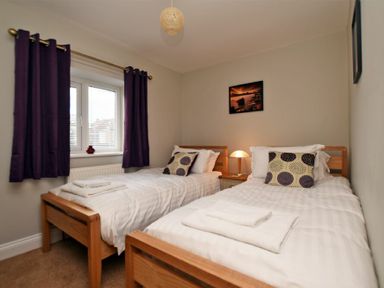 A bedroom with two single beds white bedding and patterned pillows a bedside table with a lamp and purple curtains at Schooner Court No1 in Alnmouth