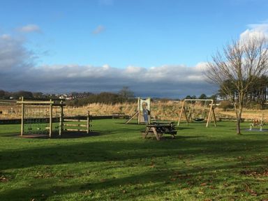A children's playground with climbing frames swings and picnic tables in a grassy field at Schooner Court No1 in Alnmouth