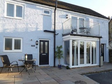 The exterior of a two storey house with a black door, windows, a small balcony with a table and chairs, and outdoor seating at Schooner Court No1 in Alnmouth