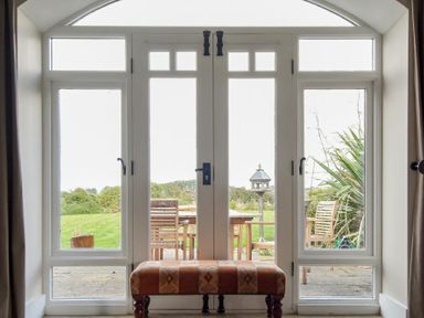 A room with a cushioned bench in front of glass doors leading to a garden with wooden chairs and a bird feeder at Sea Breeze in Newton-by-the-Sea