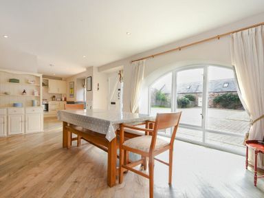 A dining area with a wooden table and chairs next to large arched windows and an open kitchen in the background at Sea Breeze in Newton-by-the-Sea