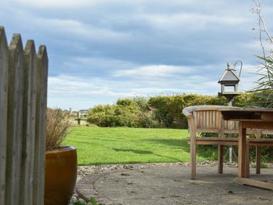 A garden with wooden bench and table on a stone patio with green lawn and shrubs at Sea Breeze in Newton-by-the-Sea