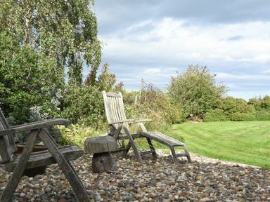 A garden area with wooden chairs on a pebble surface and grass with bushes at Sea Breeze in Newton-by-the-Sea