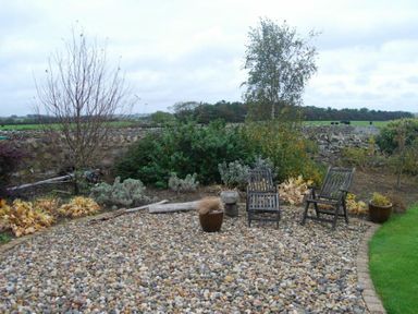 A garden area with two wooden chairs and pebble ground near bushes and trees at Sea Breeze in Newton-by-the-Sea