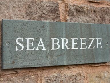 A stone wall with a green rectangular sign that reads sea breeze attached to it at Sea Breeze in Newton-by-the-Sea