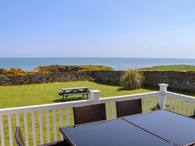 An outdoor patio with wicker chairs and a glass table overlooking a grassy yard with a picnic bench and stone wall by the sea at Seaview Howick in Craster