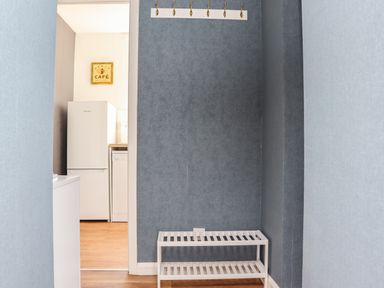 A narrow hallway with a white shoe rack and six brass hooks hanging on a dark blue wall with a view into a kitchen with a white refrigerator and dishwasher at Seaview Howick in Craster