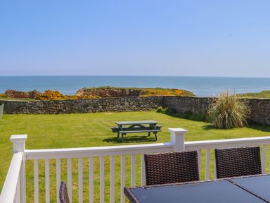A patio with a glass table and wicker chairs overlooking a grassy yard with a picnic bench and stone wall facing the sea at Seaview Howick in Craster