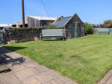 A garden with green grass benches a stone building and a stone wall at Seaview Howick in Craster