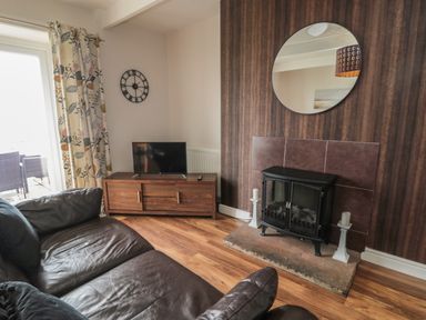 A living room with a leather sofa a wooden TV stand with a television a wall clock patterned curtains a fireplace with candles and a round mirror on the wall at Seaview Howick in Craster
